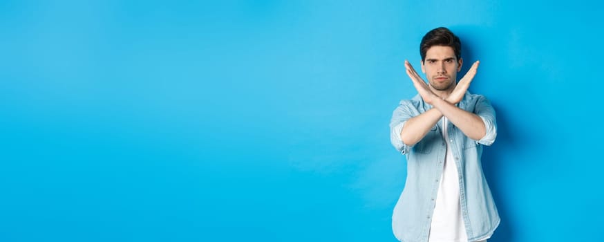 Displeased man frowning and saying no, making cross sign, tell to stop, standing against blue background.