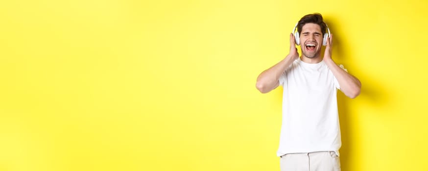 Happy guy listening music in new headphones, buying earphones on black friday, standing over yellow background.