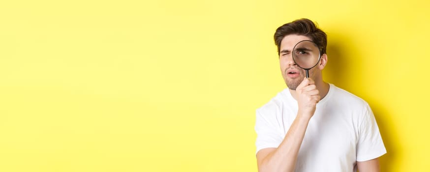 Man looking through magnifying glass with serious and thoughtful look, searching or investigating, standing over yellow background.