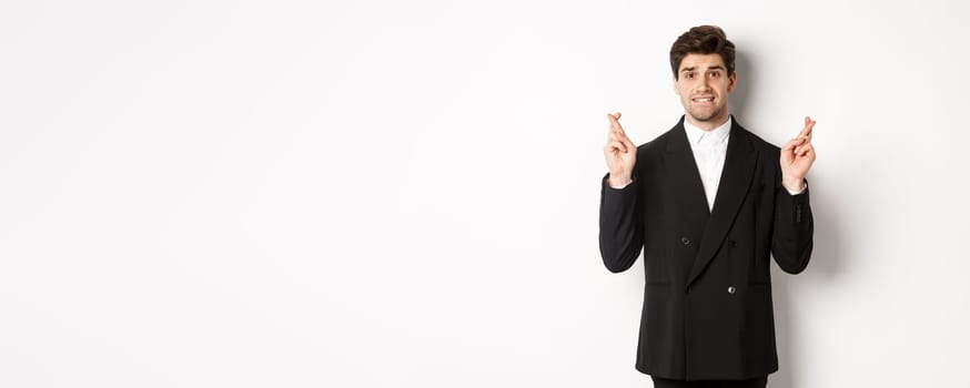 Nervous businessman in black suit crossing fingers, biting lip and making a wish, waiting for news, standing over white background hopeful.