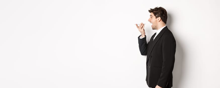 Profile shot of handsome businessman in black suit talking on speakerphone, smiling and looking happy, recording voice message, standing over white background.