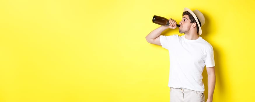Concept of tourism and vacation. Man drinking wine from bottle on holidays, standing against yellow background. Copy space