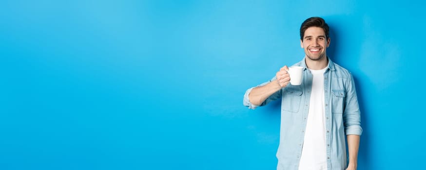 Smiling bearded man holding mug and drinking coffee, standing against blue background.