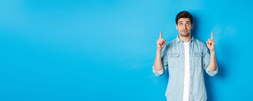 Portrait of displeased and skeptical male model pointing fingers up, looking at something unpleasant, standing against blue background.