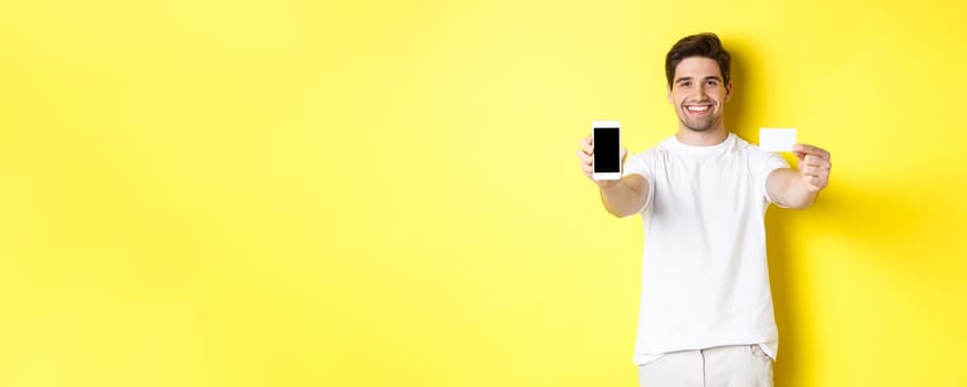 Handsome caucasian male model showing smartphone screen and credit card, concept of mobile banking and online shopping, yellow background.