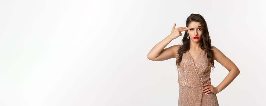 Celebration and party concept. Arrogant woman in elegant dress making finger gun at her head and looking bothered, standing annoyed over white background.