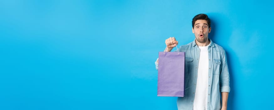 Concept of shopping, holidays and lifestyle. Handsome surprised guy holding paper bag from shop and looking amazed, standing over blue background.