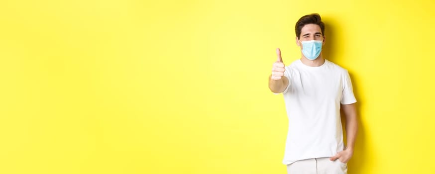 Concept of coronavirus, pandemic and social distancing. Confident young man in medical mask showing thumbs up and winking, yellow background.