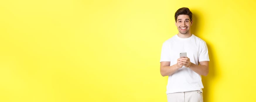 Man smiling and looking happy after reading promo offer on smartphone, standing against yellow background in white t-shirt.