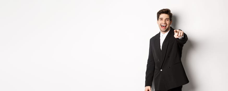 Concept of new year party, celebration and lifestyle. Portrait of handsome stylish man in black suit, smiling and pointing finger at camera, standing over white background.