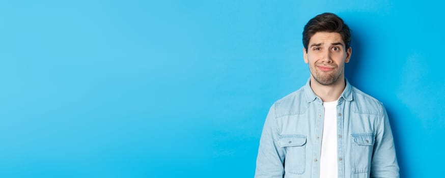 Close-up of skeptical and awkward guy smirking, feeling uncomfortable, standing over blue background.