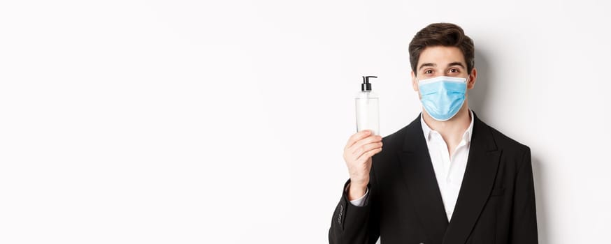 Concept of covid-19, business and social distancing. Close-up of handsome man in trendy suit and medical mask, showing hand sanitizer, standing against white background.