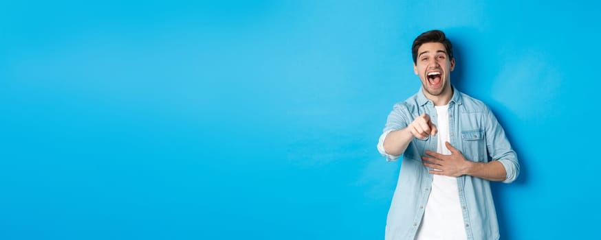 Adult man in casual outfit laughing out loud and pointing at you, looking at something funny, chuckling while standing against blue background.