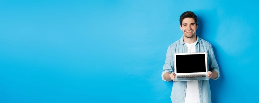 Handsome young man introduce product on laptop screen, showing computer and smiling, standing over blue background.