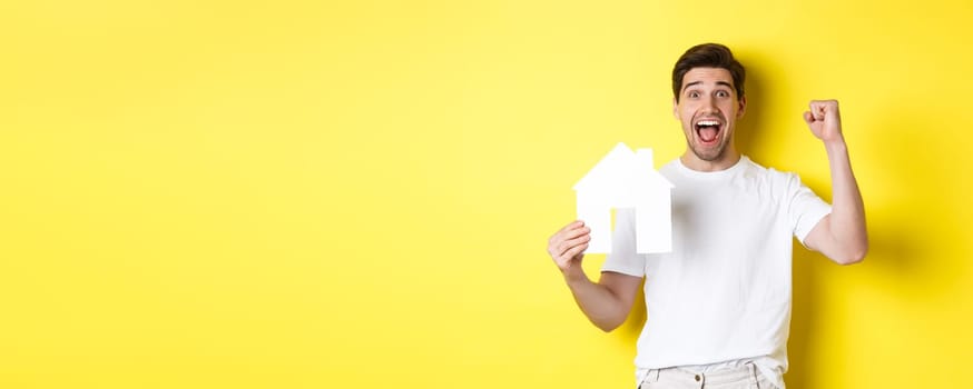Real estate concept. Cheerful man showing paper house model and making fist pump, paid mortgage, yellow background.