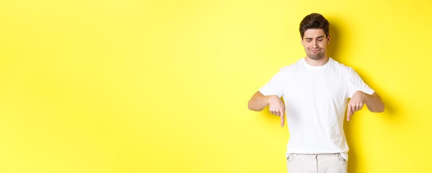 Skeptical young man in white t-shirt, pointing and looking down upset, disapprove and dislike product, standing over yellow background.