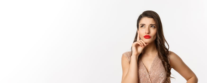 Concept of New Year celebration and winter holidays. Close-up of elegant woman with red lips and dress, looking at upper left corner and thinking, standing over white background.