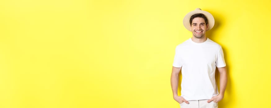 Young handsome guy tourist looking happy, wearing straw hat for travelling, standing against yellow background. Copy space