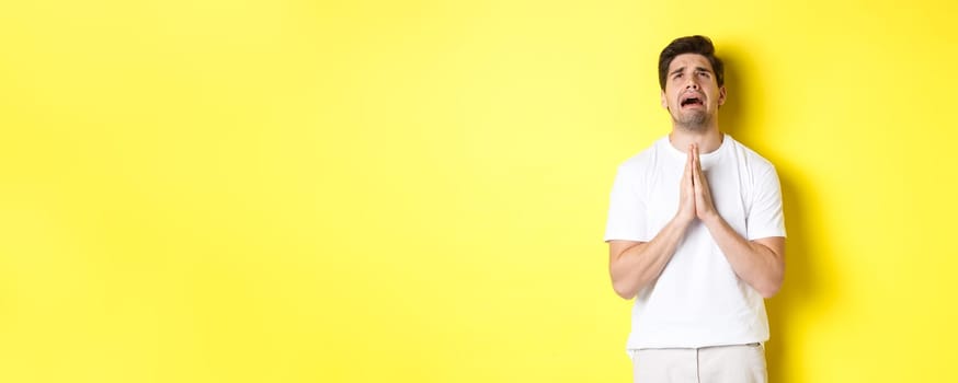 Desperate man pleading God, holding hands in pray and looking up distressed, standing over yellow background. Copy space