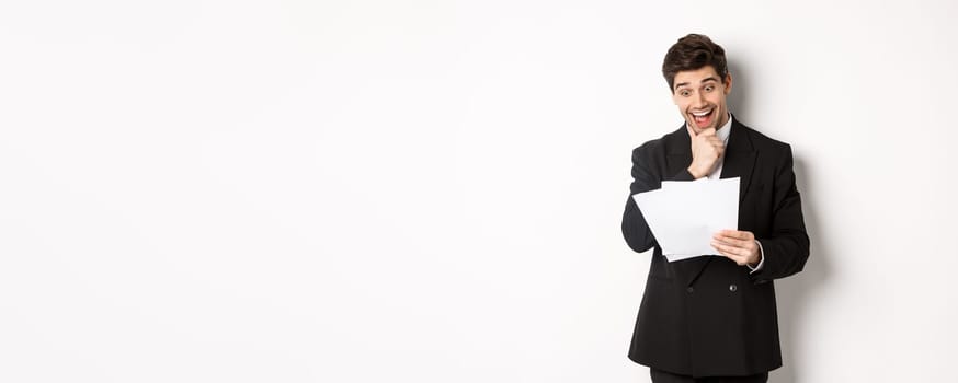 Portrait of handsome businessman looking excited at documents, working, standing against white background in black suit.