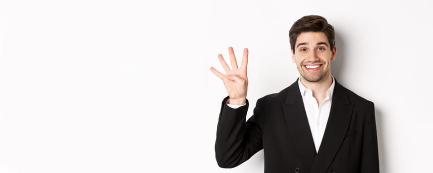Close-up of handsome businessman in black suit, smiling amazed, showing number four, standing over white background.
