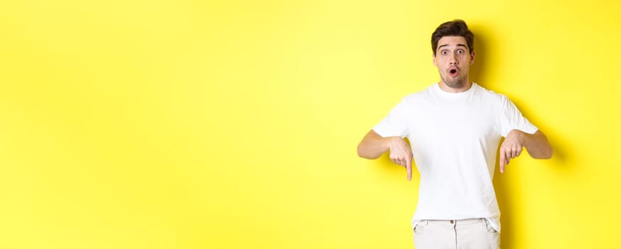 Image of impressed guy pointing fingers down, gasping amazed and looking at camera, standing over yellow background.