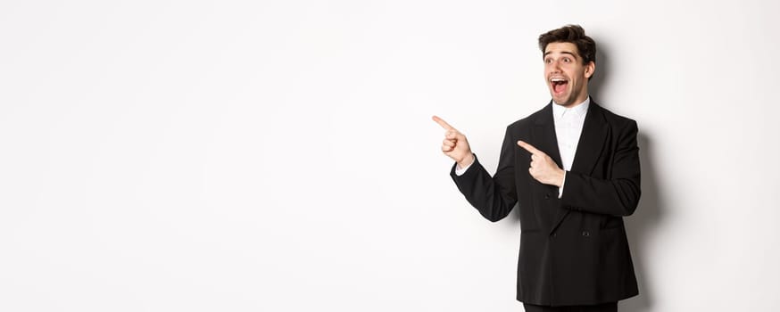 Portrait of amazed and happy handsome man in black suit, pointing and looking left impressed, showing awesome christmas promo, standing over white background.