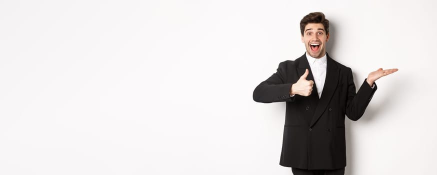 Portrait of hadnsome bearded man in formal suit, showing thumb-up and holding product in hand over white copy space, recommending product, standing over white background.