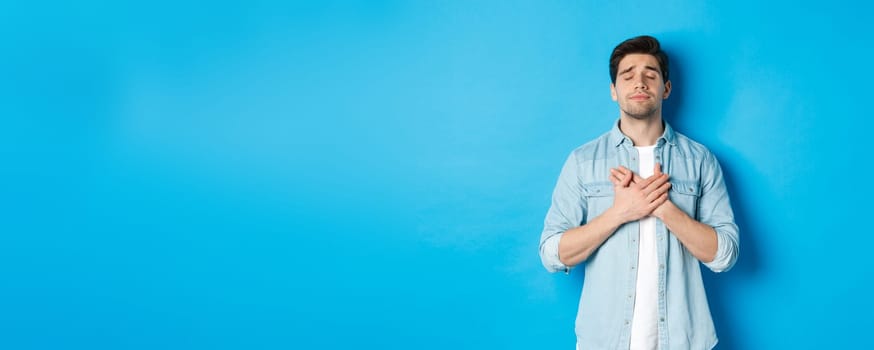 Portrait of dreamy and nostalgic guy in casual outfit, holding hands on heart and close eyes, daydreaming against blue background.