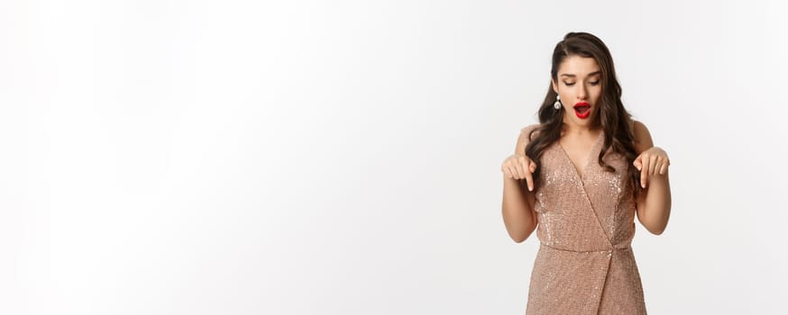 Amazed glamour girl in party dress, looking and pointing down amazed, showing christmas advertisement, standing over white background.