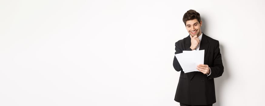 Image of handsome businessman in black suit, looking pleased at documents, reading report and smiling, standing against white background.