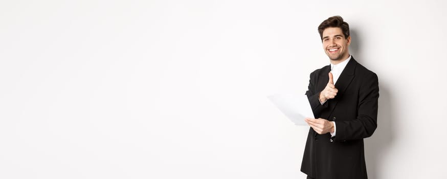 Image of handsome businessman in black suit, holding document and pointing finger at camera, praising good job, standing against white background.