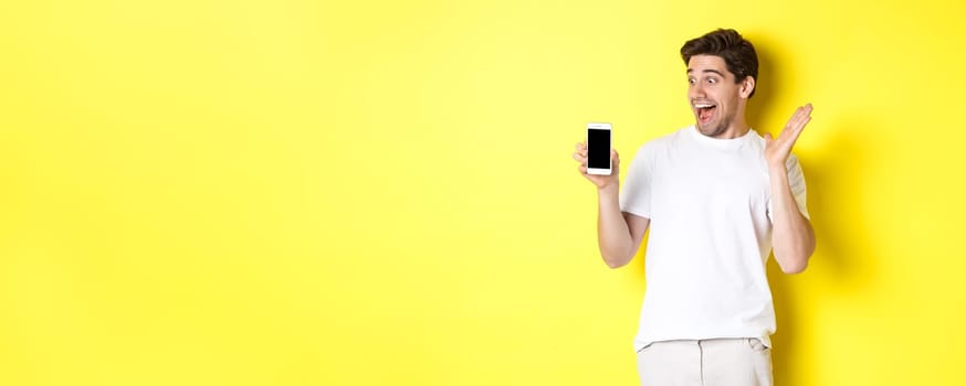 Image of amazed guy looking at mobile phone screen with surprised face, standing excited against yellow background.