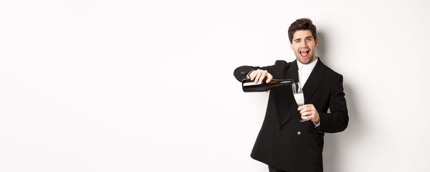 Handsome man in trendy suit pouring glass of champagne, celebrating christmas, smiling amazed and having fun, standing against white background.