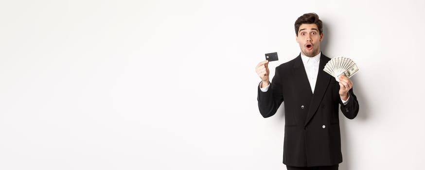 Portrait of surprised handsome man i suit, showing credit card with money, standing against white background.