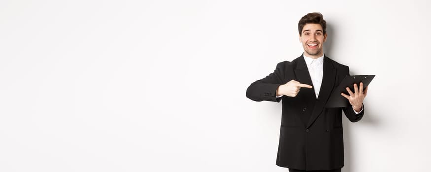 Image of handsome smiling businessman in black suit, pointing finger at clipboard with documents, standing against white background.