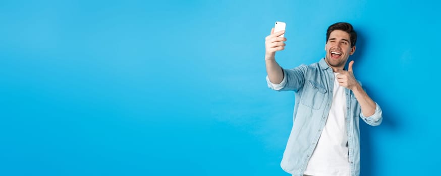 Handsome modern guy taking selfie on smartphone and pointing finger gun at mobile camera, winking cheeky, standing against blue background.