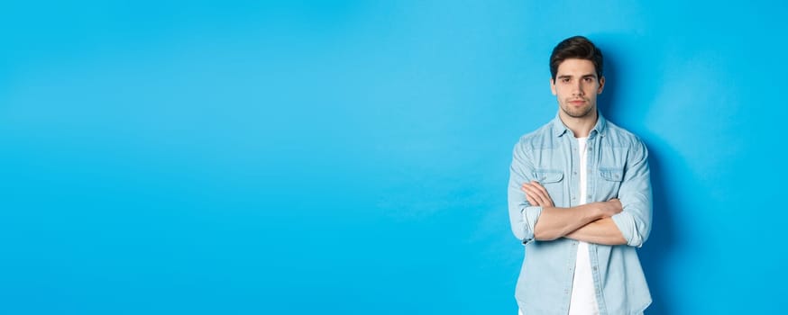 Image of handsome caucasian man in casual outfit, looking serious and confident, standing against blue background.