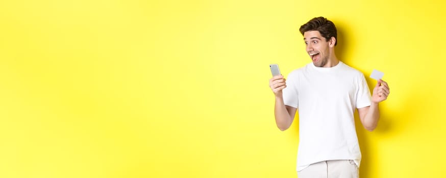 Surprised guy holding smartphone and credit card, online shopping on black friday, standing over yellow background.