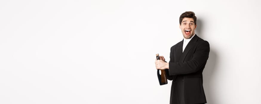 Portrait of attractive man in black suit, winking at camera and opening bottle of champagne, celebrating new year, standing against white background.