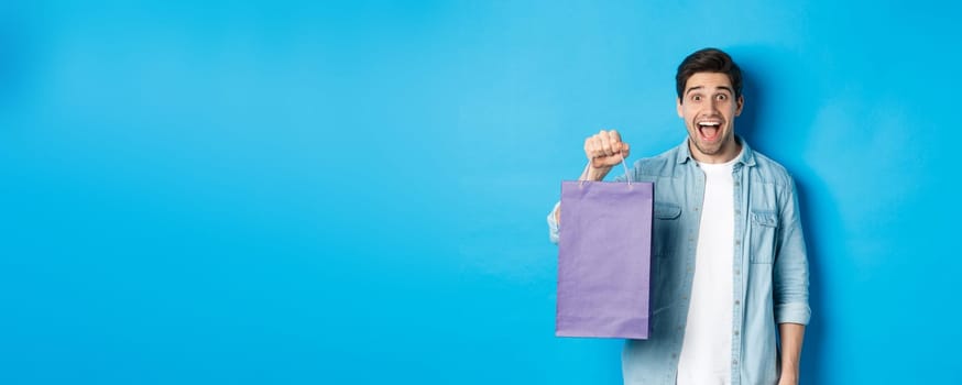 Concept of shopping, holidays and lifestyle. Excited handsome guy holding paper bag with gift and looking happy, standing over blue background.