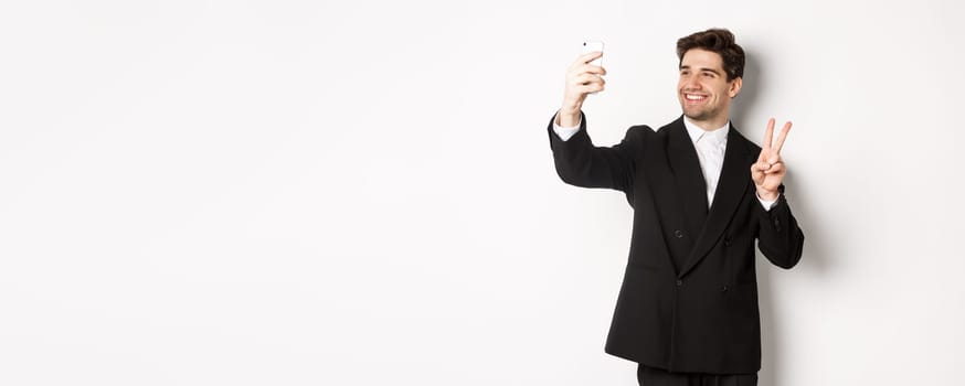 Portrait of good-looking man taking selfie on new year party, wearing suit, taking photo on smartphone and showing peace sign, standing against white background.
