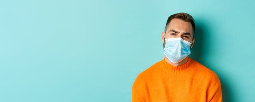 Covid-19, social distancing and quarantine concept. Upset man in face mask complaining, looking displeased and tired, standing against light blue background.