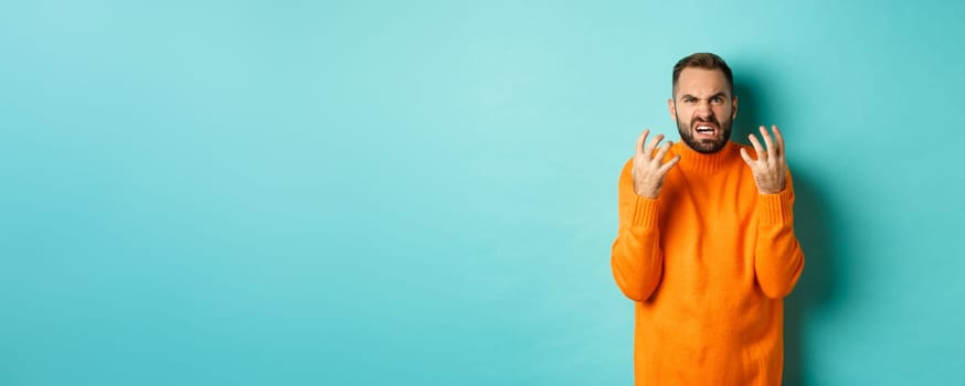 Image of angry man looking disappointed and mad, shaking hands and grimacing with disdain, standing distressed against light blue background.