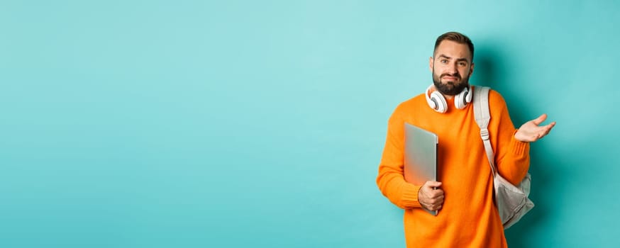 Image of troubled young man with headphones and backpack, shrugging confused and holding laptop, standing over light blue background.