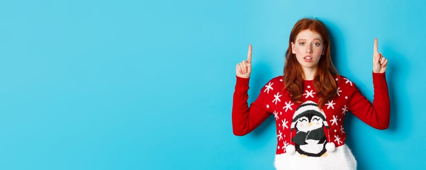 Merry Christmas. Skeptical and unamused redhead girl pointing fingers up, showing logo with reluctant face, standing over blue background.