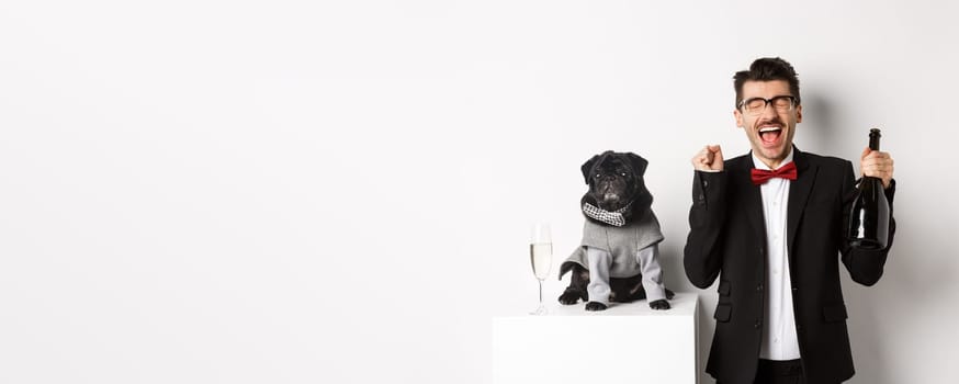 Pets, winter holidays and New Year concept. Happy young man celebrating Christmas with cute black dog wearing party costume, holding bottle champagne, white background.