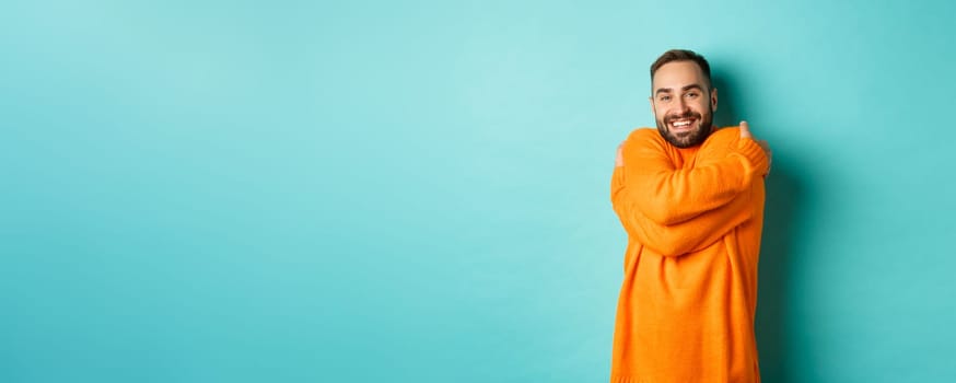 Happy man feeling comfortable, wearing warm sweater and hugging himself, smiling satisfied, standing over light blue background.