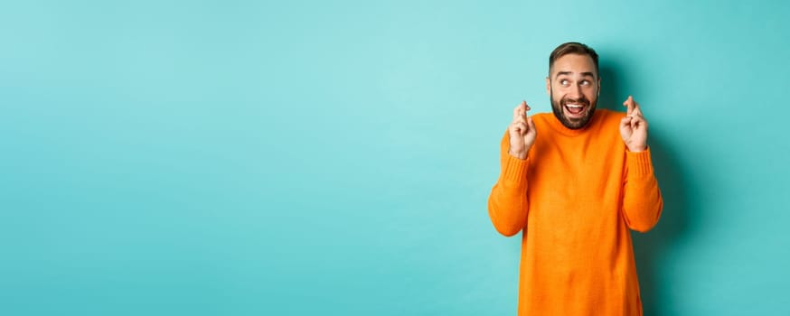 Excited man expecting something with anticipation, hold fingers crossed and smiling, making wish, standing over light blue background.