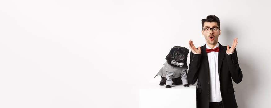 Animals, party and celebration concept. Image of dog owner and cute pug in costumes suits staring surprised at camera, reacting on promo offer, white background.